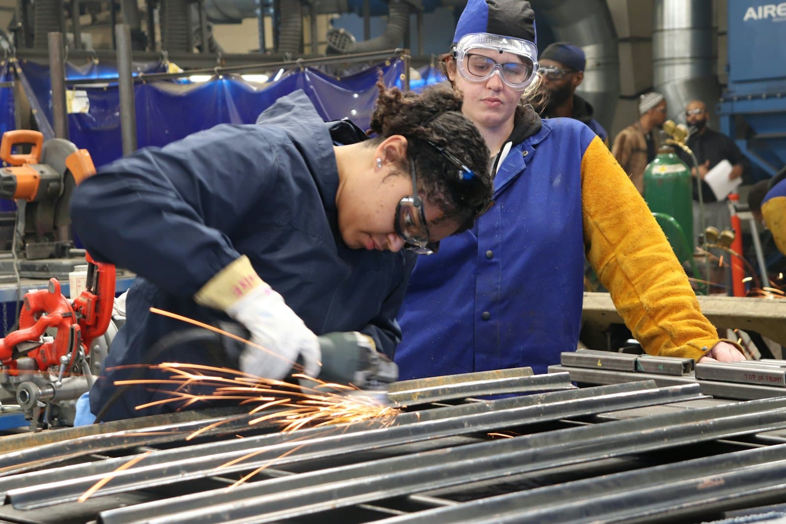 Two trainees in safety gear working at a metal workshop bench.